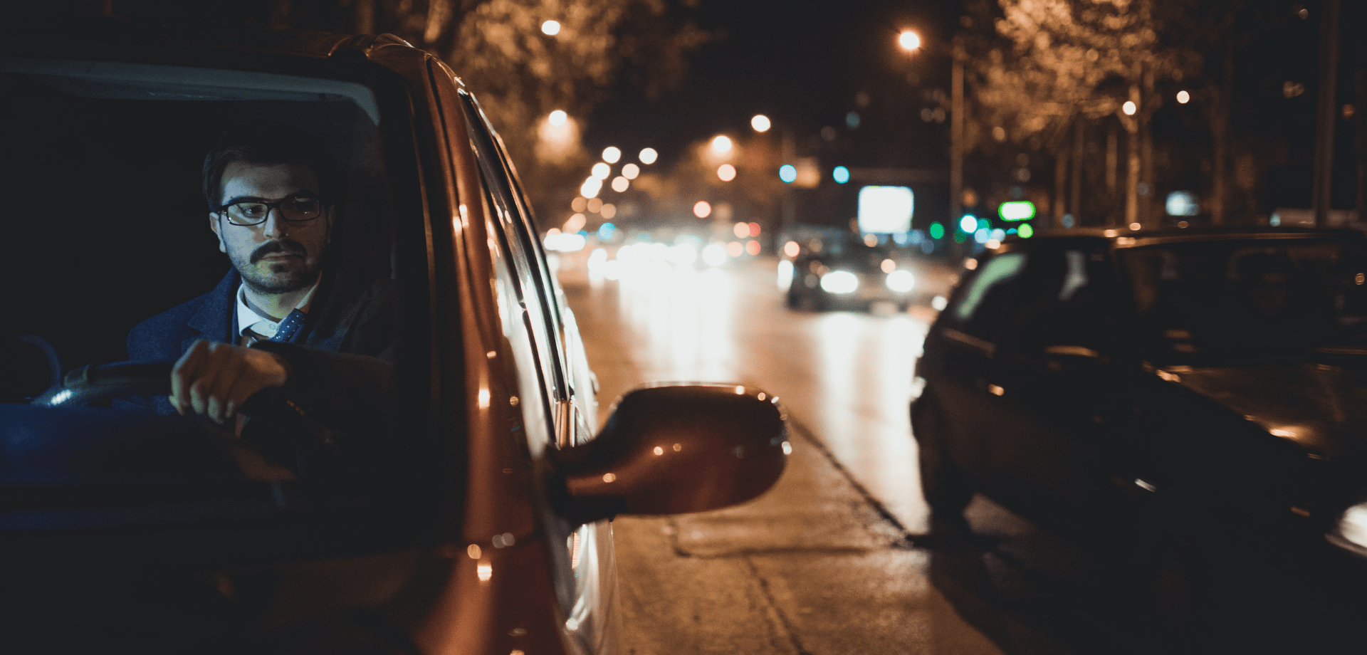 Image of a man wearing glasses driving a car at night time.